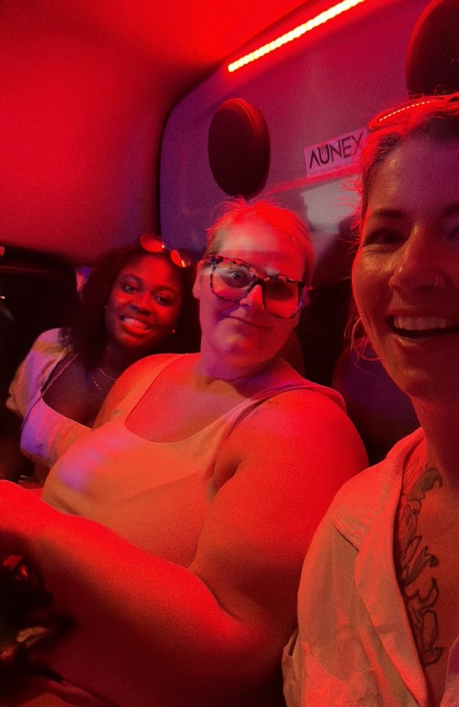 three women sitting together in a back bus seat glowing in a red overhead light that looks like a nightclub