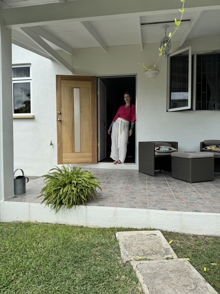 a woman wearing a hot pink shirt and white pants standing in an open doorway on the back covered porch of a home in barbados with green plants and wicker seating