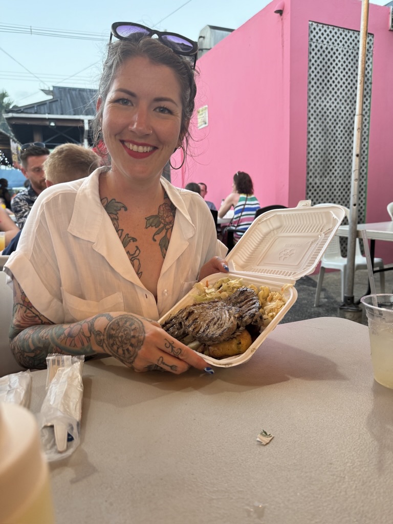 a woman seated at an outdoor eating area holding up a takeout container of grilled food